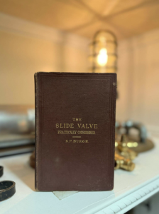 A dark brown vintage book titled 'The Slide Valve' standing upright on a white mantle with a blurred lamp in the background.