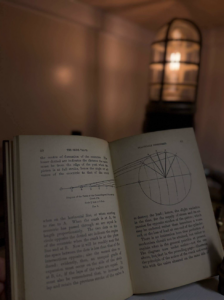 Open book showing technical diagrams and dense text, with a circular wheel diagram on the right page; a warm lamp blurs in the background.