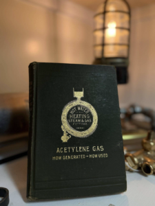Black vintage book cover reading 'Acetylene Gas: How Generated—How Used' with brass fittings nearby and a warm lamp in the background.