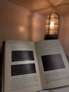 Open book with technical diagrams and text, focused on left pages, warm lamp glow in the background behind the pages.