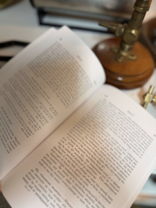 Open book on a desk, with a vintage brass lamp in the background and a small brass fitting nearby.
