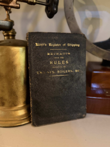 Old black hardcover book titled 'Lloyd's Register of Shipping' in gold lettering, containing extracts from the rules on engines and boilers, set beside a brass lamp.