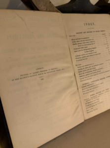 Open antique book showing an index on the right page and publication info on the left; a dark metal object rests along the top edge of the pages.