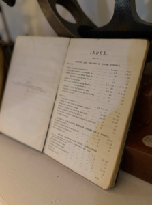 Open vintage book showing an 'INDEX' page with table of contents about engines and boilers of steam vessels, set on a stand or surface with a dark machine part in the background.
