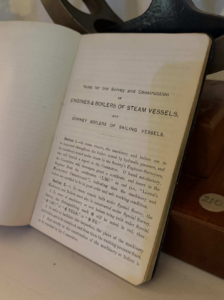 Open vintage manual showing title: 'Rules for the Survey and Construction of Engines & Boilers of Steam Vessels' on a desk edge.
