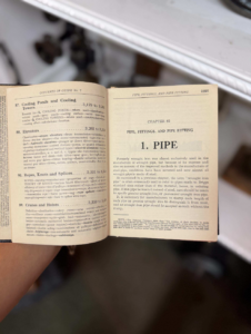 Open vintage technical book held by a hand, showing Chapter 82: PIPE, FITTINGS, AND PIPE FITTING.
