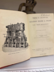 Open vintage technical book showing a detailed line drawing of triple expansion engines and machinery on the left page.