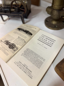 Open vintage book showing a chapter titled 'The Modern Steam Car and Its Background' on a white table, with antique machinery in the background.