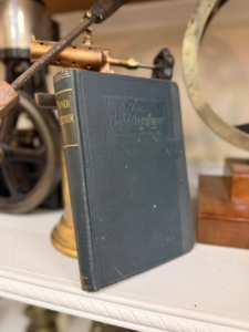 Antique dark-blue hardcover book titled 'The Power Plant' standing upright on a white shelf with vintage machinery in the background.