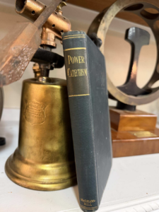 Close-up of a brass bell beside a dark blue hardcover book titled 'Power Catechism' on a shelf.