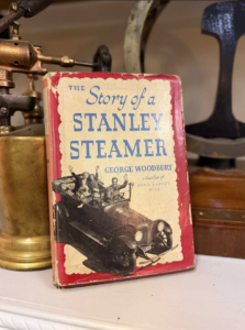 Cover of a vintage book titled The Story of a Stanley Steamer by George Woodbury, featuring a black antique car illustration on a red-bordered cover. It leans against brass machinery on a shelf.
