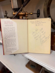 Open book showing a page titled 'The Story of a Stanley Steamer' with a typed article on the left and handwritten notes on the right, against antique machinery in the background.