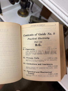 Open older book showing Contents of Guide No. 8: Practical Electricity, DC, chapters 91–94 (Electricity, Primary Cells, Conductors and Insulators, Electrical and Mechanical Energy) with a sink in the background.