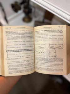 Open vintage technical book about transformers, showing diagrams and dense text across a two-page spread, held by a hand.