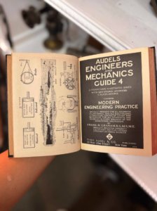 A person holds an open vintage engineering book with left-hand diagrams and a bold cover page reading 'AUDels ENGINEERS AND MECHANICS GUIDE 4' on the right side.