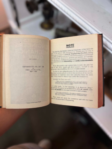 Open vintage book held by a hand, right page labeled NOTE with dense paragraph text visible.
