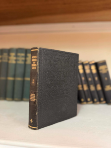 Small black textured hardcover book standing upright on a shelf, titled 'Audels Engineers and Mechanics Guide' (volume 2) with similar volumes blurred in the background.