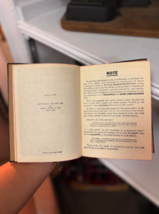 Open antique book held in a person's hand, showing the right-hand page with a bold 'NOTE' heading and dense small print.