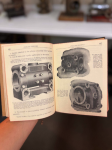 Open vintage manual showing engine diagrams, held by a person with a blurred background.