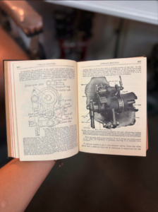 Open book showing engine diagrams and a photo of a mechanical component, held by a person in a dim background.