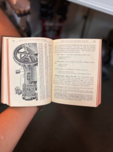 Person holding an open technical manual showing a detailed engine diagram on the left and dense text on the right page.