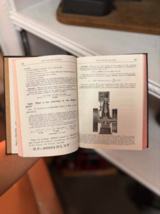 Open book showing a vintage steam engine manual, with text on left page and a technical illustration on the right page, held by a hand above a desk.