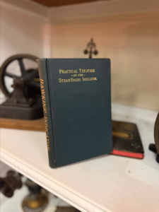 Blue hardcover antique book titled 'Practical Treatise on the Steam Engine Indicator' with gold lettering, standing upright on a white shelf beside a vintage wheel in the background.