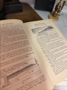 Open book with diagrams on a desk, title 'Maxims and Instructions' visible, brass lamp in background