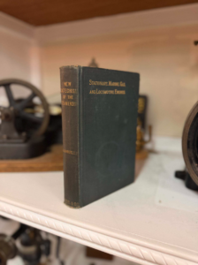 Dark green antique hardcover book standing upright on a white shelf with blurred vintage machinery in the background, left of center.
