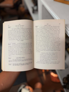 Open book held by a person, pages filled with dense text, background blurred (indoor setting). It shows two facing pages with small typewritten text on aged paper, hands visible at bottom edge of book.