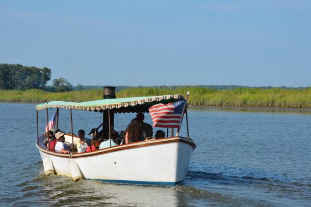 People ride on a small boat with an American flag, floating on calm water with reeds in the background.