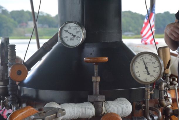 Close-up of a vintage steam boiler with two pressure gauges and valve handles on a boat, American flag visible in the background.