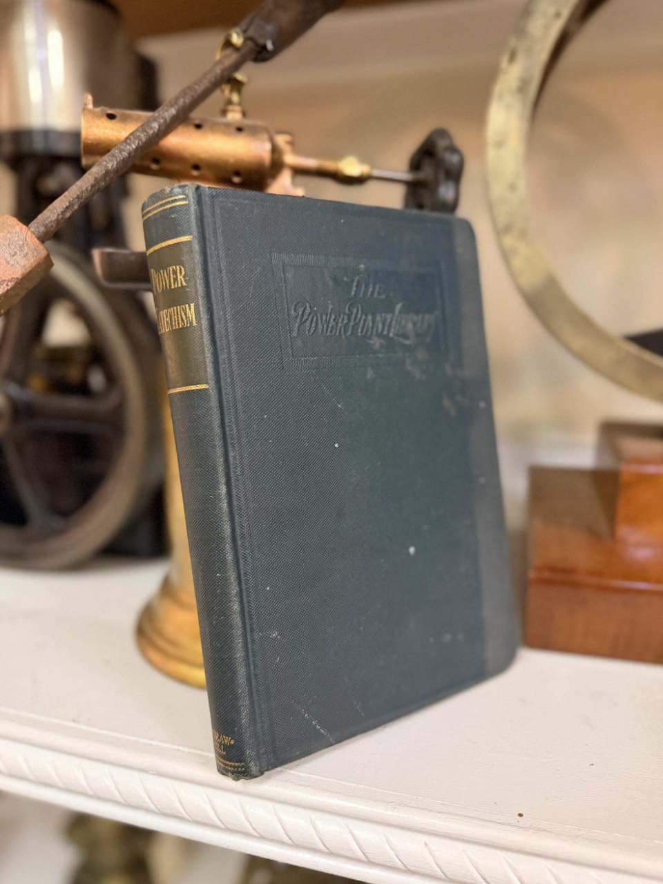 Antique dark-blue hardcover book titled 'The Power Plant' standing upright on a white shelf with vintage machinery in the background.