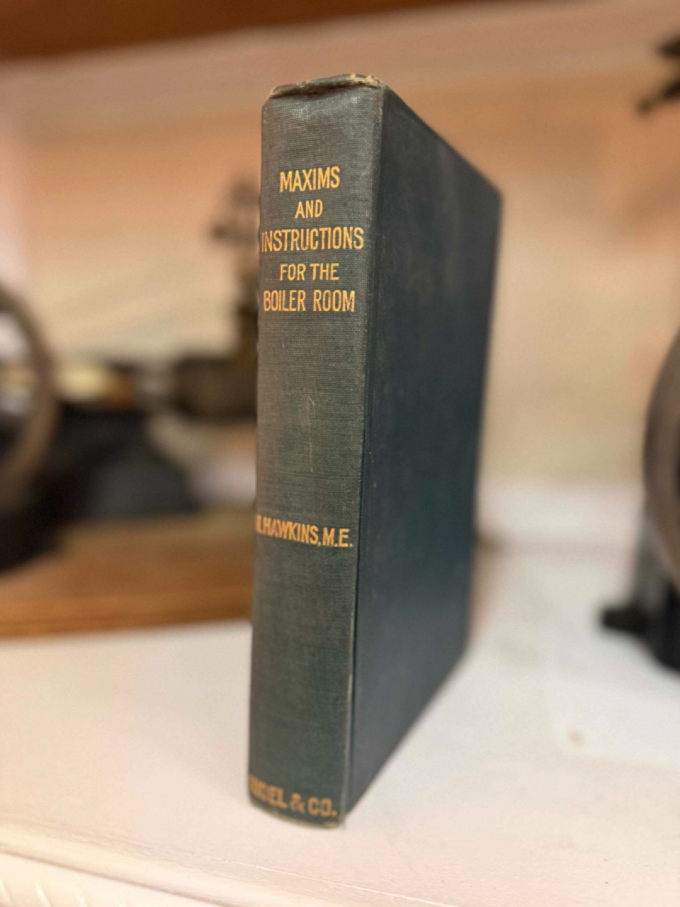 Dark green hardcover book standing upright on a white surface, gold lettering reads 'Maxims and Instructions for the Boiler Room' with author name at the bottom of the spine.