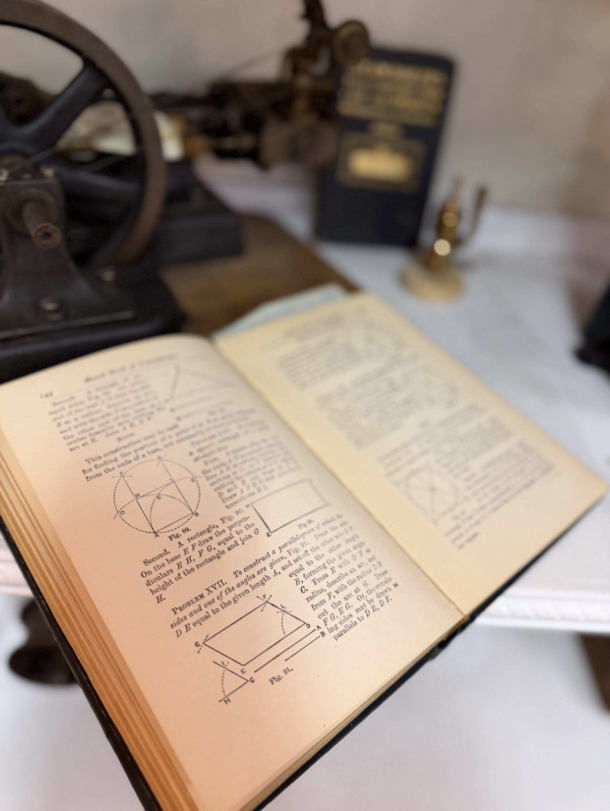 Open book with geometric diagrams and dense text on a wooden table; a vintage machine and a second book blur in the background.