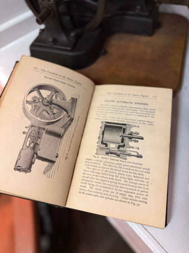 Open vintage technical manual showing engine diagrams on yellowed pages; a metal machine sits in the background.