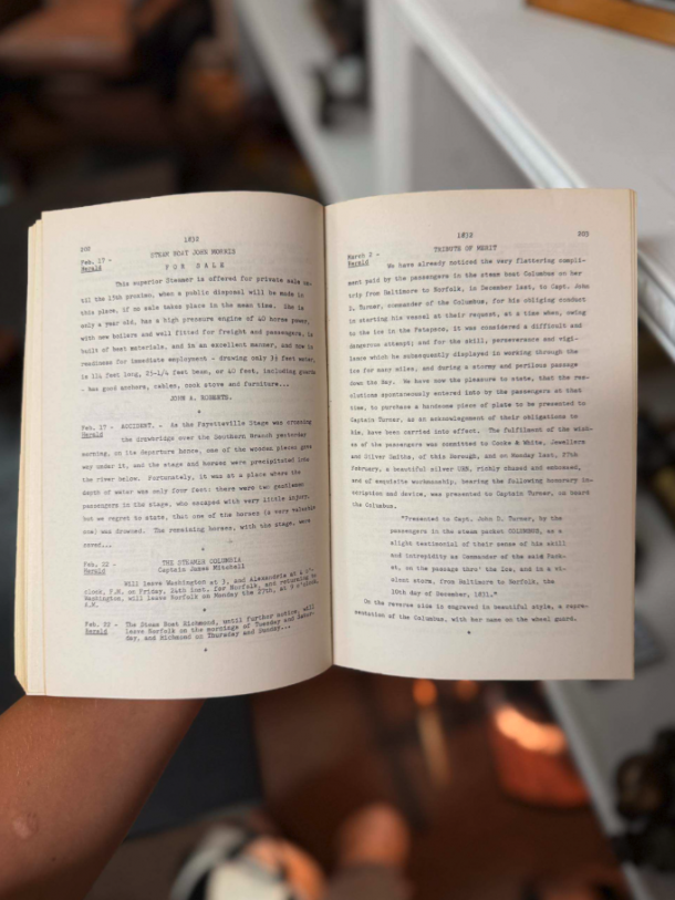 Open book held by a person, pages filled with dense text, background blurred (indoor setting). It shows two facing pages with small typewritten text on aged paper, hands visible at bottom edge of book.