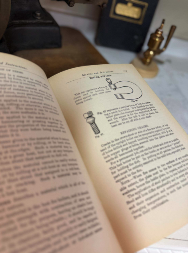 Open vintage instruction book showing mechanical diagrams and dense text on cream pages, with brass tools in the background on a wooden desk.