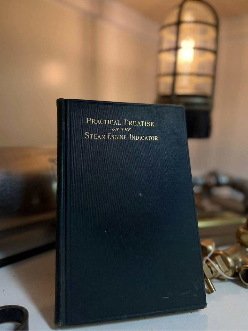 Black hardcover book titled 'Practical Treatise on the Steam Engine Indicator' resting on a table with a warm lamp glowing in the background.