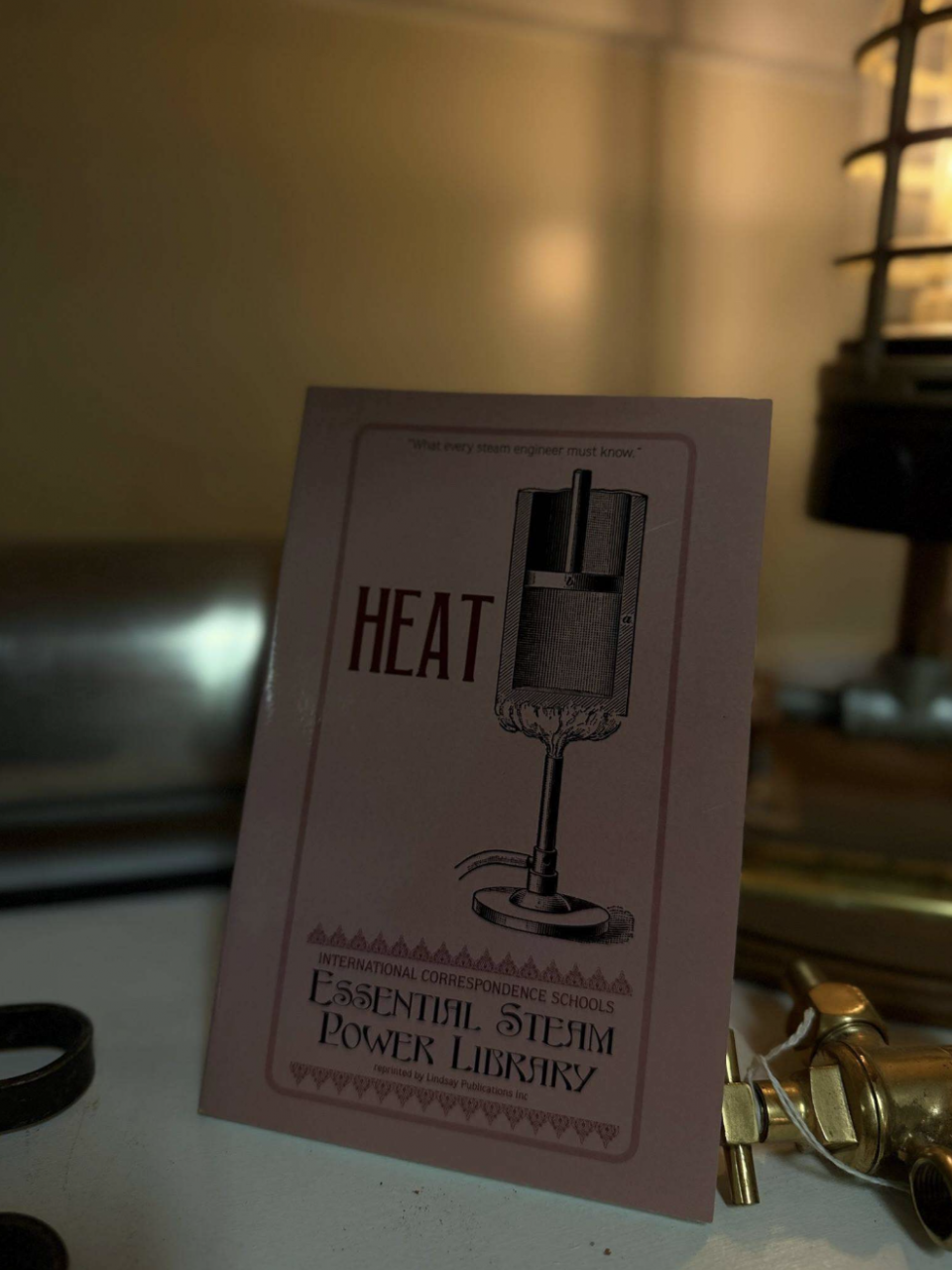 A pink card with a vintage illustration of a steam power device and the word 'HEAT' in red, leaning on a desk with blurred background objects nearby.